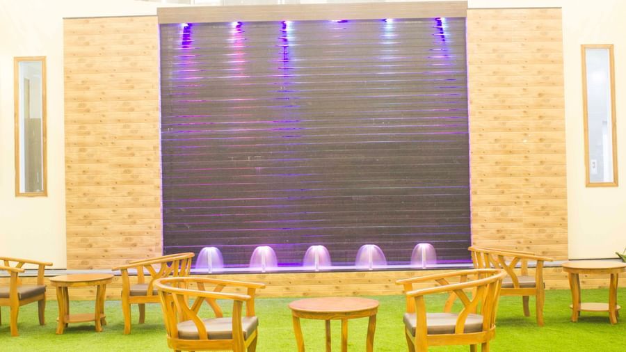An indoor area with wooden chairs and tables on green artificial grass, facing a water wall | Daksh The Madhuvan Suites