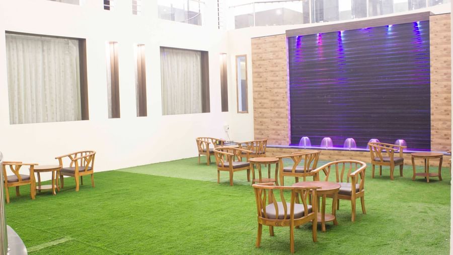 An indoor area with wooden chairs and tables on green artificial grass, facing a water wall | Daksh The Madhuvan Suites