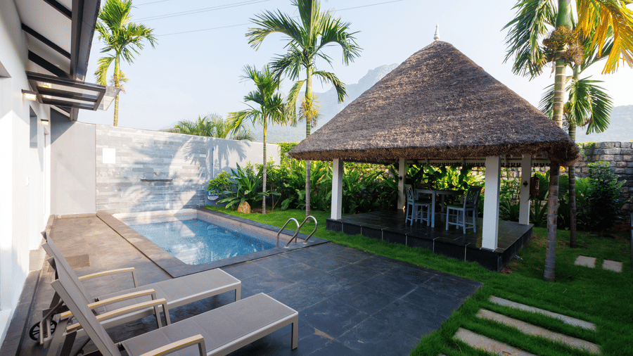 The outdoor area view of the Grand Pool Villa with loungers, a plunge pool, a canopy with a thatched roof and chairs inside on a manicured garden inside Darza Resorts.