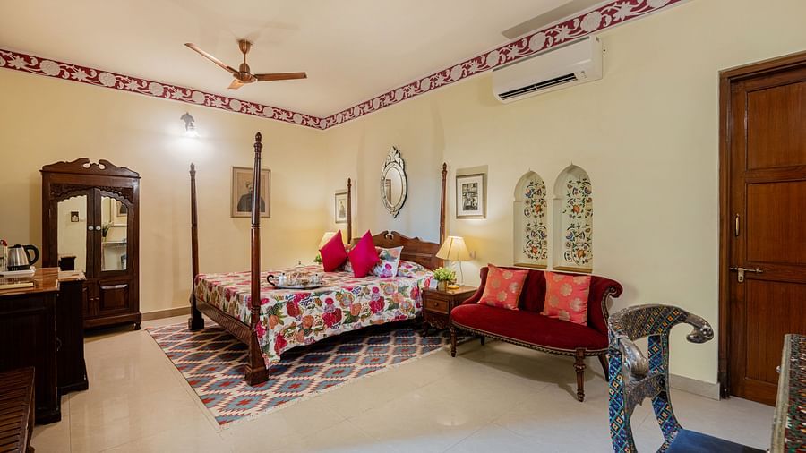A bedroom with a four-poster bed, patterned bedding, a red sofa, and ornate trim along the ceiling | Dera Rawatsar, Jaipur