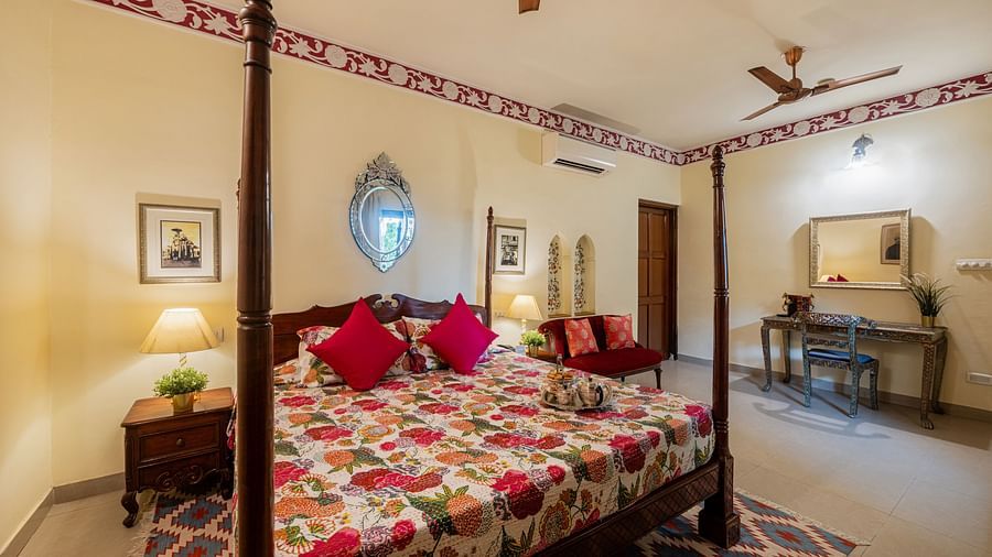 A comfortable bedroom at Dera Rawatsar in Jaipur includes a four-poster bed, a seating area, a dressing table, and decorative wall accents.