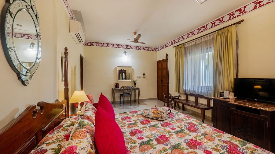 A bedroom with a patterned bedspread, red cushion, ornate mirror, ceiling fan, and decorative trim along the ceiling | Dera Rawatsar, Jaipur