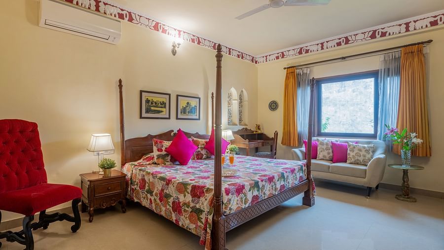 A bedroom with a four-poster bed, patterned bedding, pink cushions, a red armchair, and decorative trim along the ceiling | Dera Rawatsar, Jaipur