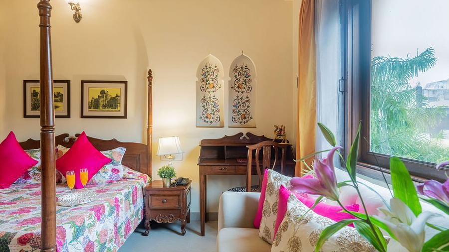 A bedroom with a four-poster bed, patterned bedding, pink cushions, a wooden desk, and a window with a view of greenery | Dera Rawatsar, Jaipur