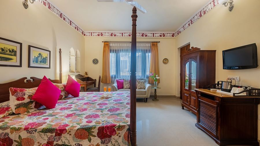 A bedroom with a four-poster bed, patterned bedding, pink cushions, a television, and decorative trim along the ceiling | Dera Rawatsar, Jaipur