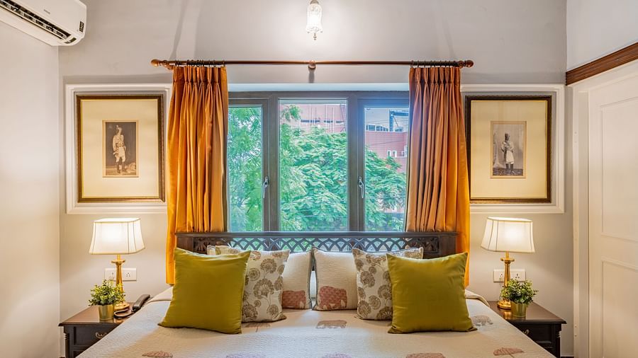 A tranquil bedroom at Dera Rawatsar in Jaipur features a comfortable bed, bedside lamps, framed artwork, and a window with vibrant curtains.