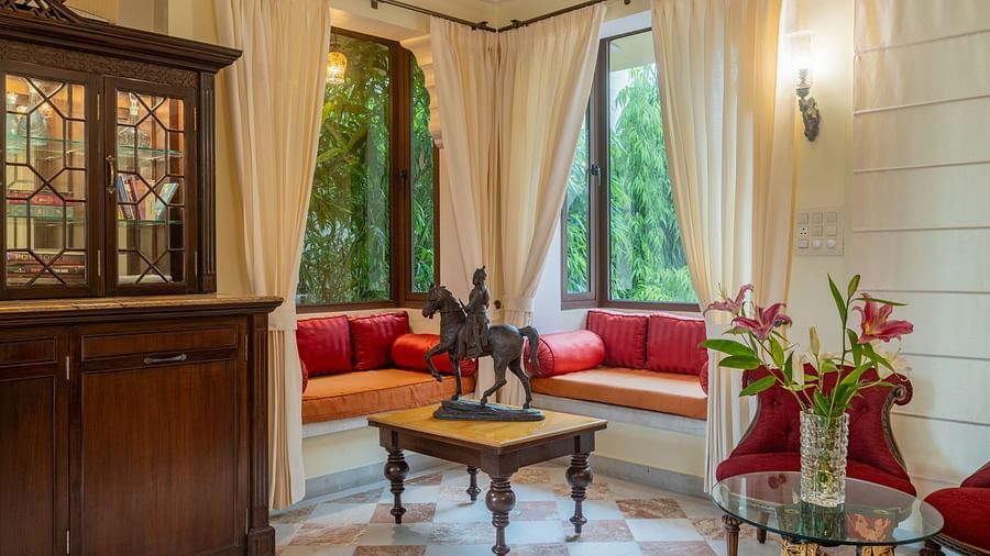 A charming sitting room at Dera Rawatsar features window seats with cushions, a wooden coffee table with a statue, and a glass-topped side table with flowers.
