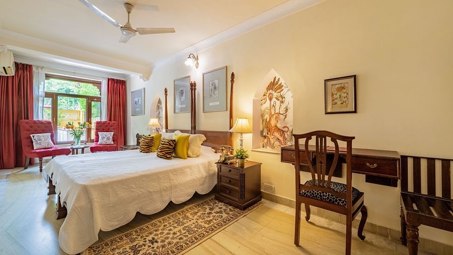 A bedroom with a four-poster bed, white linens, yellow cushions, a wooden desk, and a decorative niche in the wall | Dera Rawatsar, Jaipur