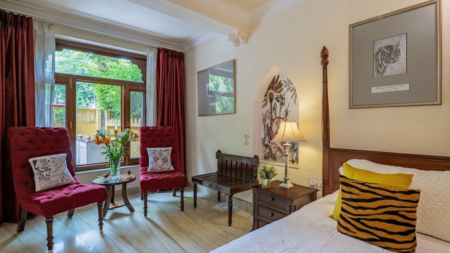 A bedroom with a four-poster bed, red armchairs, a patterned cushion, and a window with red curtains | Dera Rawatsar, Jaipur