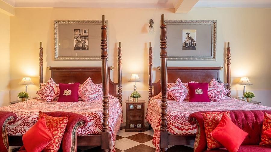 A bedroom with two four-poster beds, patterned bedding, red cushions, and matching framed artwork above each bed | Dera Rawatsar, Jaipur