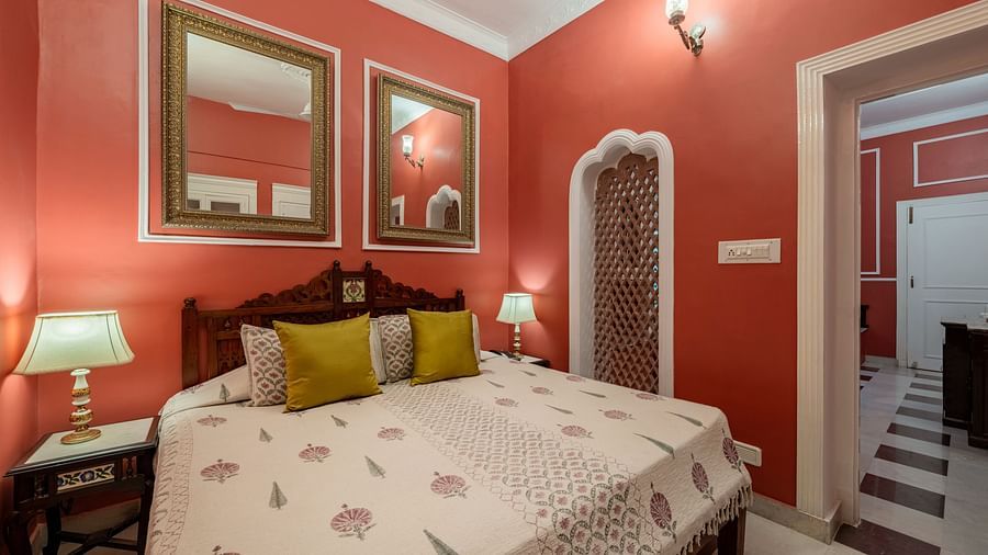 A vibrant bedroom at Dera Rawatsar, Jaipur, features a comfortable bed, framed mirrors, bedside lamps, and an ornate niche in the wall.