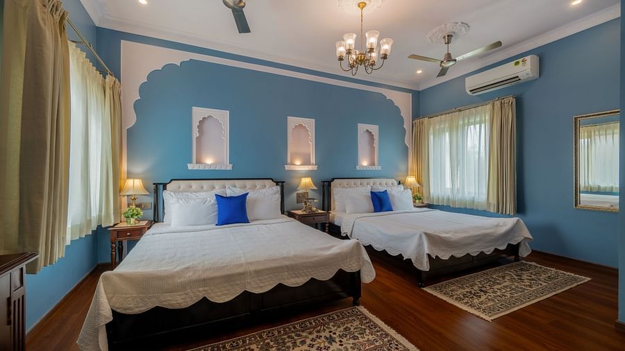A blue-walled bedroom with two twin beds, blue cushions, arched niches, ceiling fans, and sheer curtains | Dera Rawatsar, Jaipur