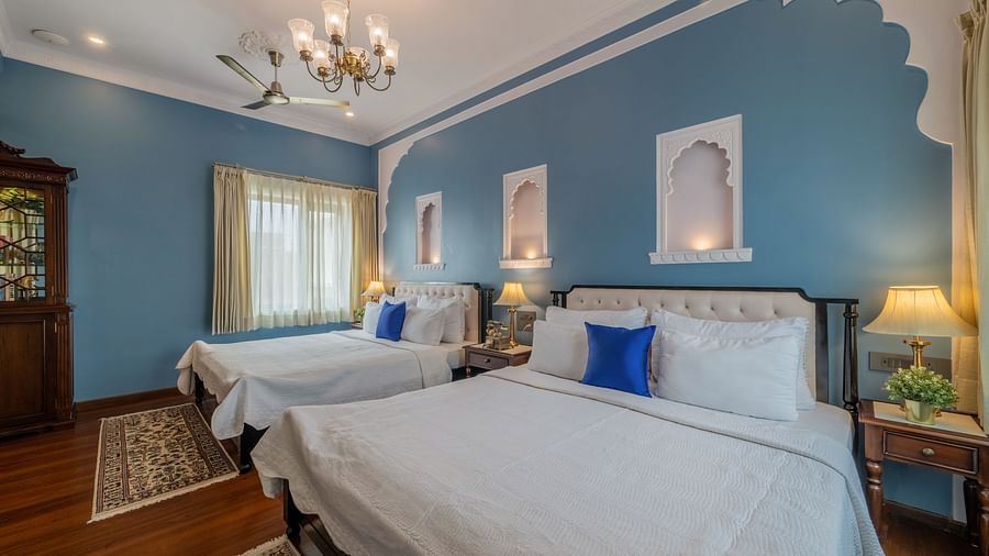 A blue-walled bedroom with two twin beds, blue cushions, arched wall niches, and a chandelier | Dera Rawatsar, Jaipur