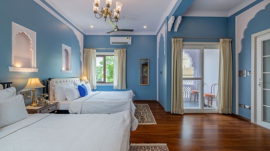 A bright and airy room at Dera Rawatsar, Jaipur, offers two comfortable beds, a balcony with seating, elegant wall details, and wooden flooring.