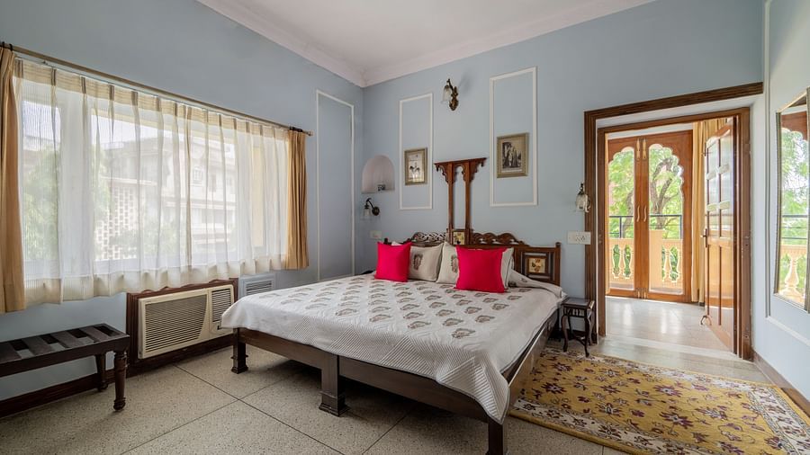 A bedroom with a light bedspread, red cushions, a wooden bed frame, and French doors leading to a balcony | Dera Rawatsar, Jaipur