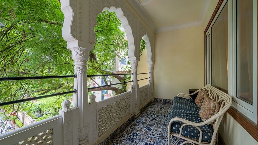 A balcony with ornate arched railings, patterned tile floor, and a wicker chair overlooking green trees | Dera Rawatsar, Jaipur