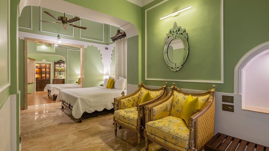 A charming suite at Dera Rawatsar, Jaipur, features two comfortable beds, stylish armchairs, an ornate mirror, and elegant archway details.