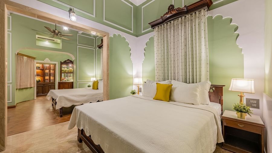 A serene suite at Dera Rawatsar, Jaipur, features a comfortable four-poster bed, an additional single bed in an adjacent area, and elegant wall paneling.