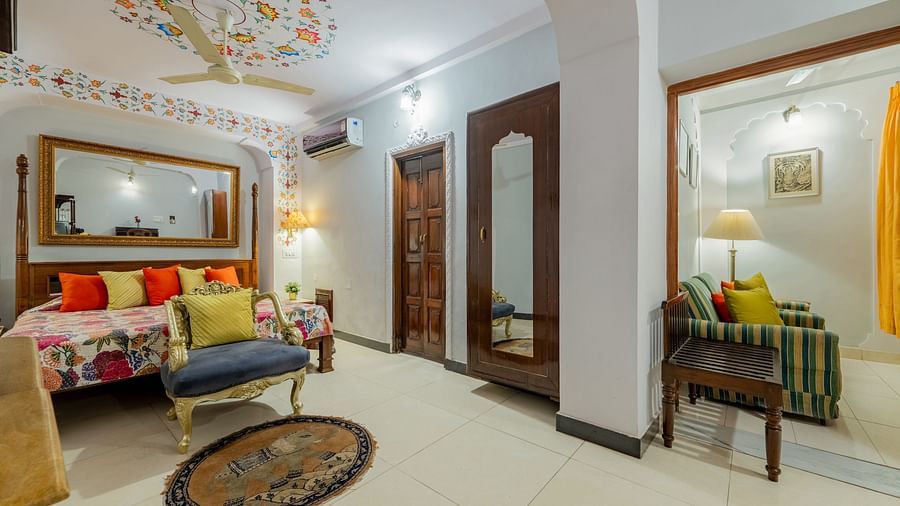 A room with a patterned ceiling, a large mirror, a bed with colorful cushions, a blue armchair, and a doorway leading to another room | Dera Rawatsar, Jaipur