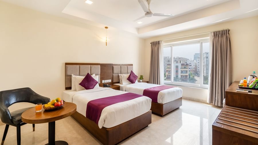 A Deluxe Room at Destino by Monday Hotels, Gachibowli, with twin beds, a small table with fruits, and a large window providing natural light.