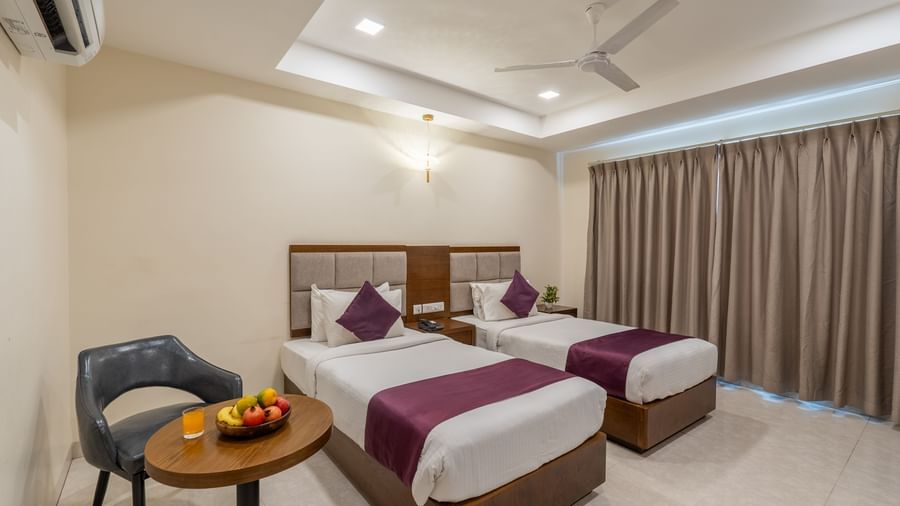 A Deluxe Room at Destino by Monday Hotels, Gachibowli, with twin beds, a small table with a fruit basket, and a window with its drapes closed.