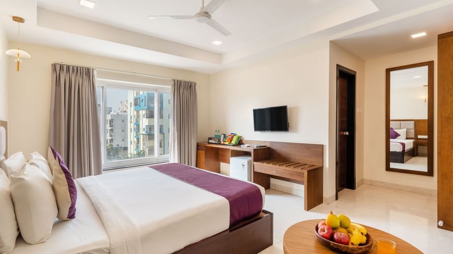 A spacious and well-lit room at Destino by Monday Hotels, Gachibowli, featuring a neatly made bed, a television, and a small round table with a fruit bowl.