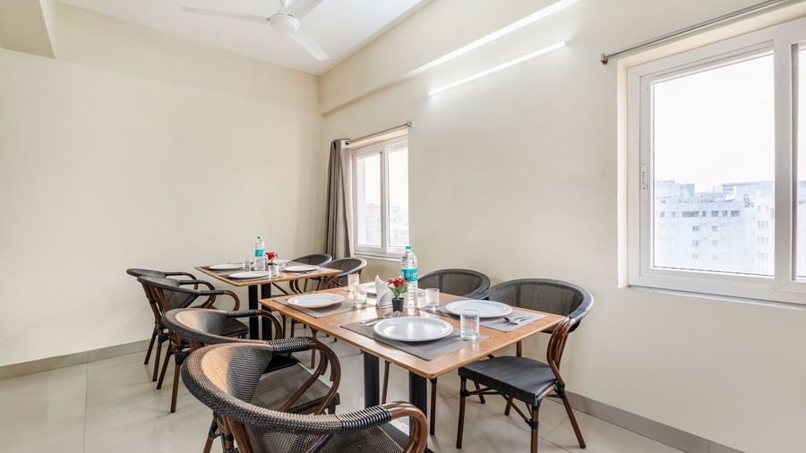 A close-up shot of a table in the dining area of Destino by Monday Hotels, Gachibowli, set with plates, glasses, and a bottle of water next to a window with grey curtains.
