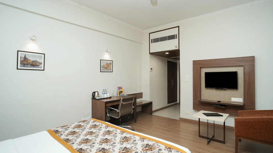 A bedroom at Diamond Hotel Varanasi features a wooden desk, a mini-fridge, and framed wall art.