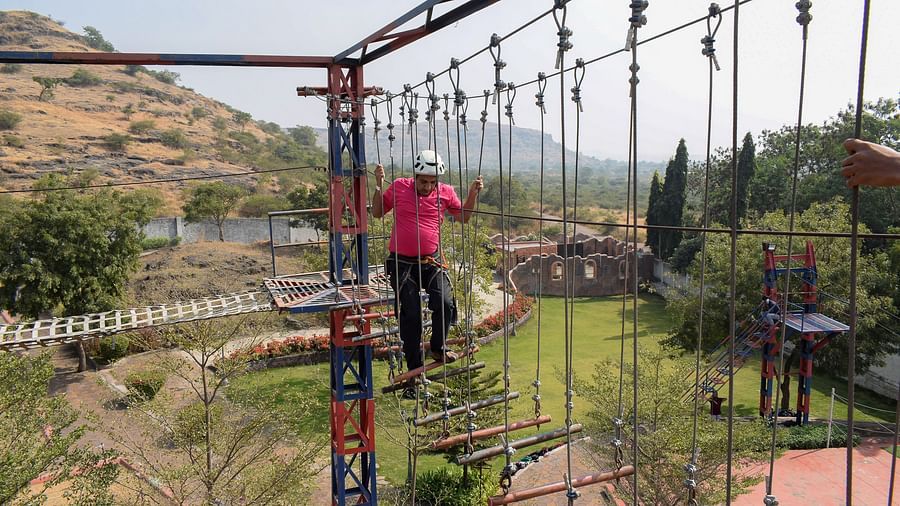 A person traversing a high rope obstacle course at an outdoor adventure park, scenic hills in the background | Diamond Parks, Pune