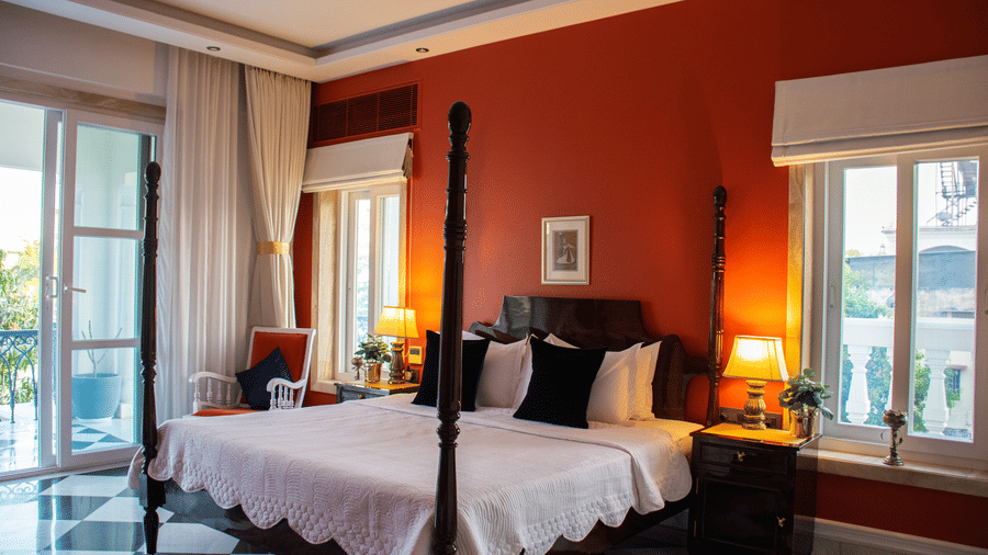 A room at Dileep Kothi, Jaipur, with a four poster double bed, side tables with night lamps, French windows, and a red wall behind the bed