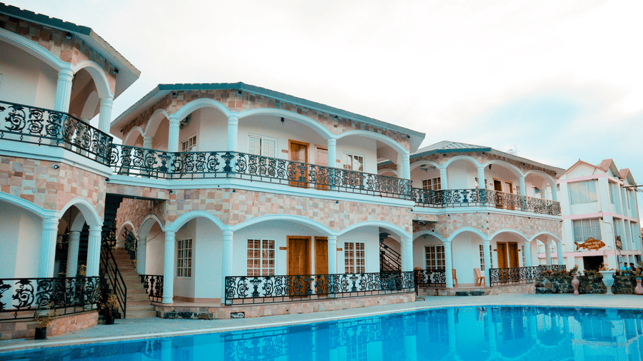 Facade of Dip Holidays in Birbhum from a different angle featuring cyan coloured building with black metal railings with a clear swimming pool in the foreground.