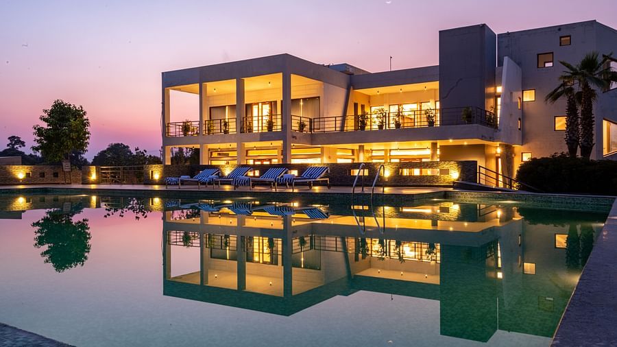 Facade view of DLS Forest Vines Resort, with warm light glowing from its windows and doors, reflected on the shimmering swimming pool beneath a clear sky.