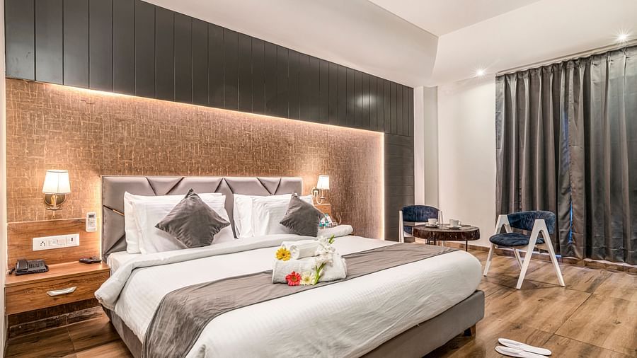 An image of the deluxe room with a bed, bed side tables, wall mounted lamps, chairs and tables, slippers and a window with grey curtains - DLS Ganga Bliss, Haridwar