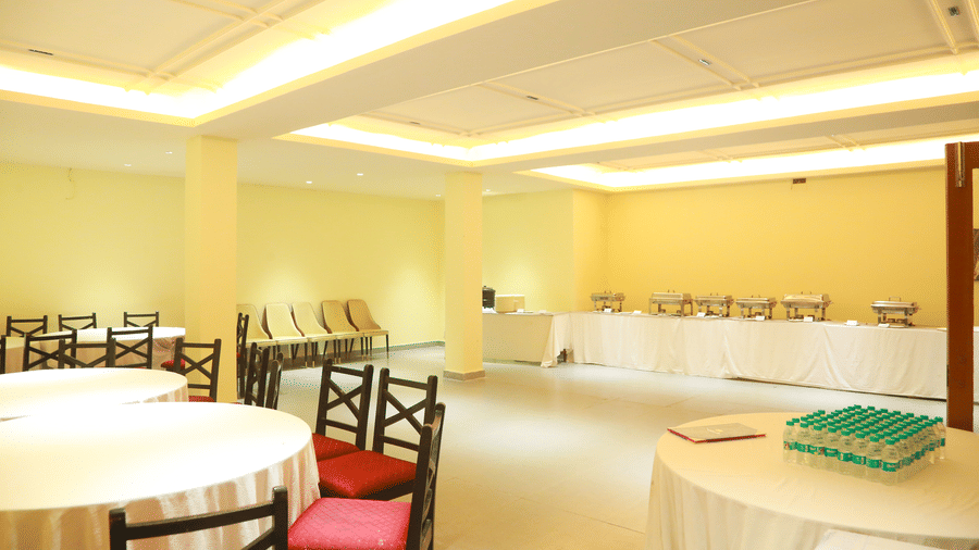 The bright Carnation banquet hall at DNC Shevaroys Resort & Spa with round tables, white linens, and a buffet station under recessed lighting.