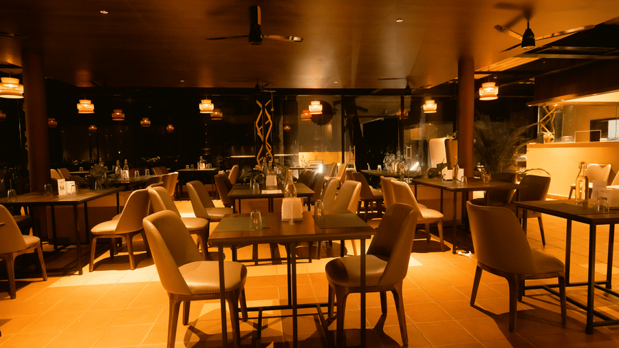 The ambiently lit restaurant at DNC Shevaroys Resort & Spa featuring dark wooden tables and upholstered chairs under warm ceiling lights.