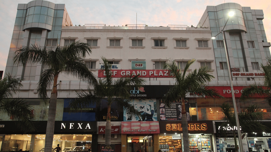 The exterior of DSF Grand Plaza, Tuticorin at dusk, showing the hotel signage above ground-floor shops, with palm trees and pedestrians on the street below.