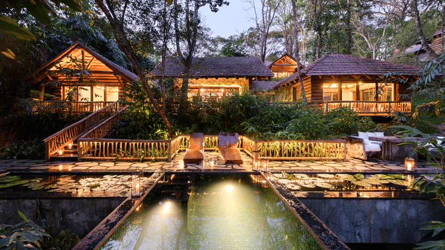 Pool and lily ponds at Estate Aana Pana at dusk, with carved loungers, candle lanterns and log buildings lit from within at Earthitects Holiday Experiences.