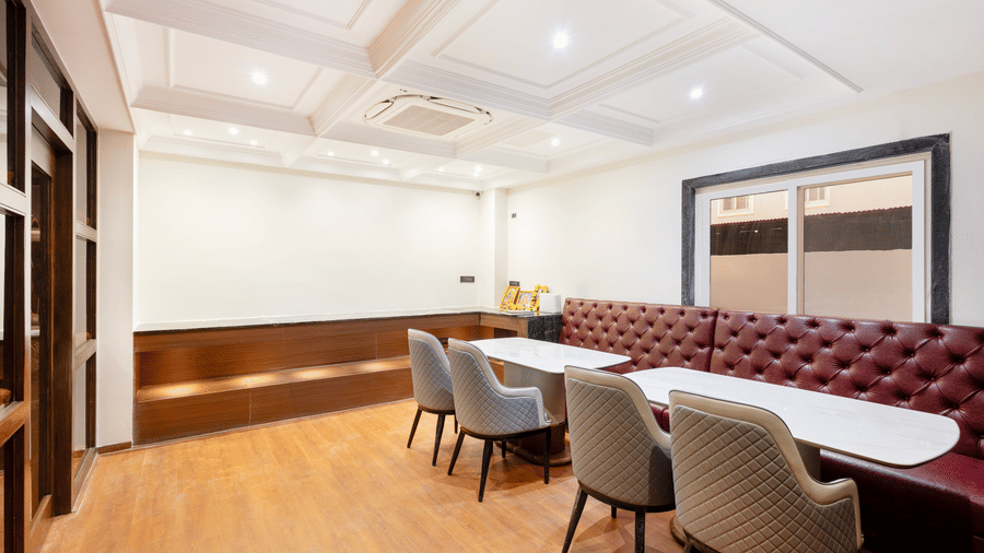 A contemporary dining area with a long, tufted maroon banquette, light wood flooring, and modern grey chairs at white tables at ELYSIUM Premier Suites, HITEC City.
