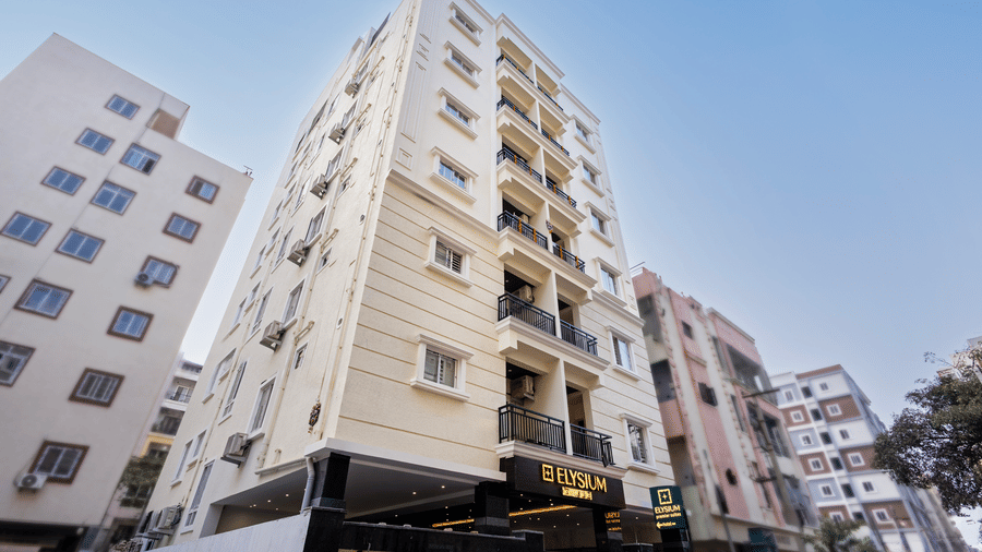 A low-angle shot of the modern, multi-storey Elysium hotel building under a clear blue sky, showing its pale facade and balcony railings at ELYSIUM Premier Suites, HITEC City.