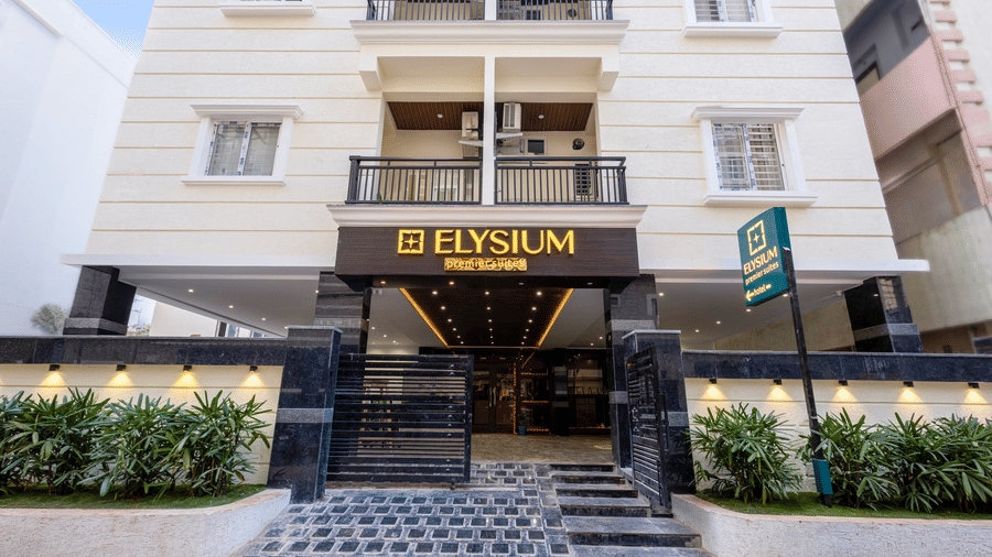 The ground-floor entrance of the Elysium hotel, featuring a dark stone-tiled ramp, green shrubs, and a prominent gold-lettered sign at ELYSIUM Premier Suites, HITEC City.