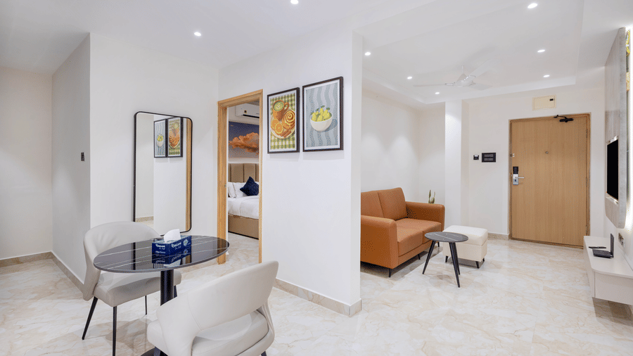 A bright, open-plan hotel suite featuring a small dining table for two, a brown leather sofa, and clean tiled flooring throughout at ELYSIUM Premier Suites, HITEC City.