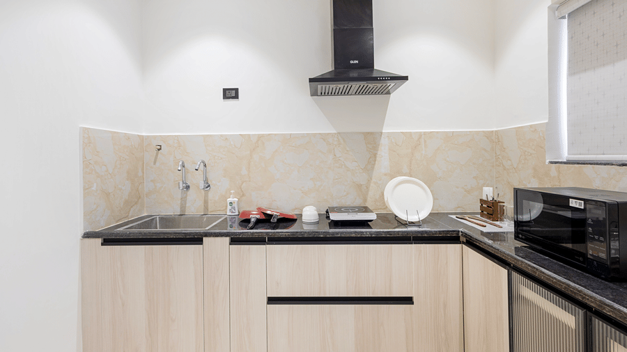 A clean, minimalist hotel kitchenette featuring a black countertop, integrated sink, and modern appliances against neutral tiled walls at ELYSIUM Premier Suites, HITEC City.