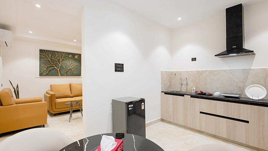 A modern open-plan hotel suite featuring a small dining table, a tan leather sofa, and a compact kitchenette with a black induction hob at ELYSIUM Premier Suites, HITEC City.