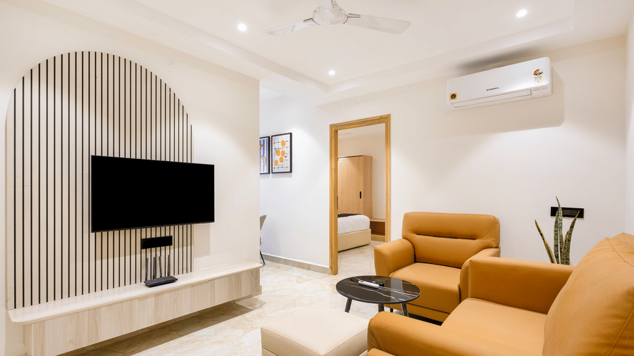 A bright living area in a hotel apartment with a wall-mounted TV, stylish wooden slat feature, tan armchairs, and polished flooring at ELYSIUM Premier Suites, HITEC City.