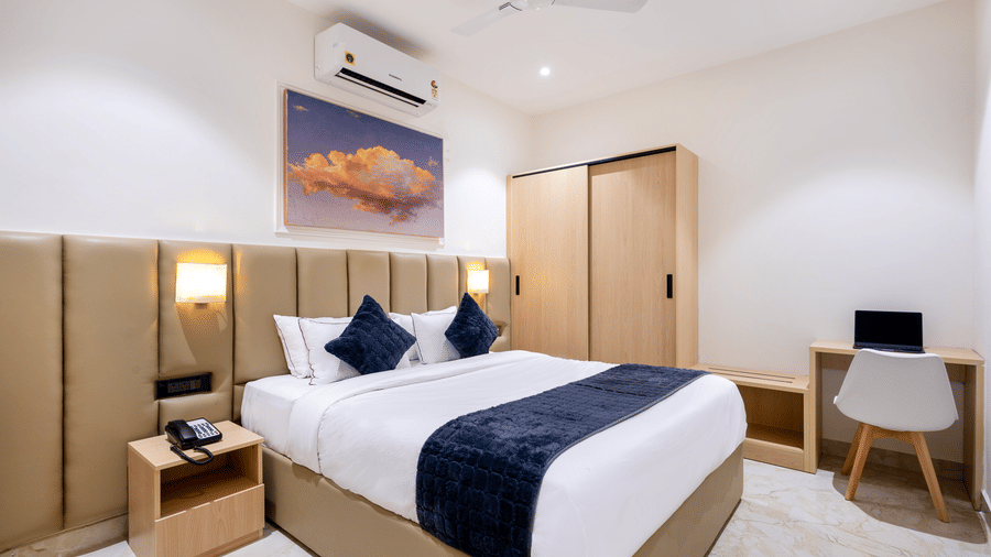 A corner view of a contemporary bedroom showing a well-lit vanity desk, a minimalist chair, and a light wood wardrobe next to the bed at ELYSIUM Premier Suites, HITEC City.
