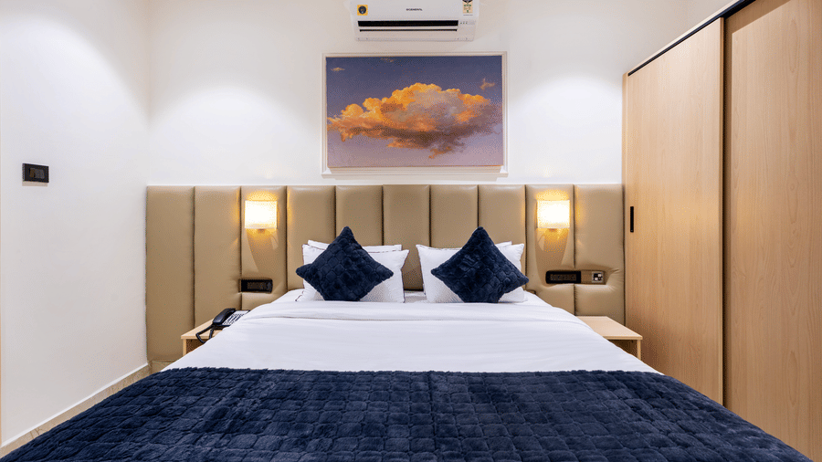 A symmetrical view of a modern hotel bedroom featuring a plush headboard, navy blue cushions, and a large sunset painting above the bed at ELYSIUM Premier Suites, HITEC City.