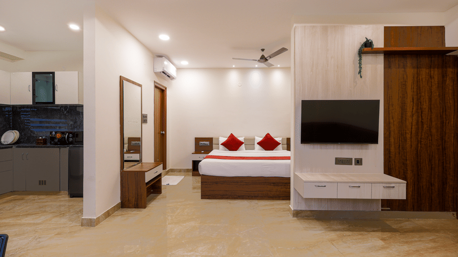 A room at Elysium Studio Suites, Madhapur, shows a bed, a wooden cabinet, and a television mounted on the wall.