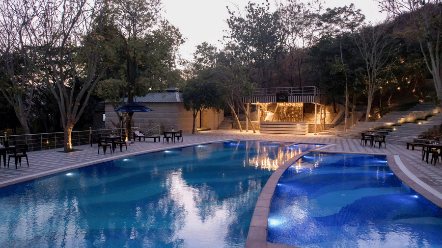 Swimming pool at Essentia Luxury Resort & Spa, Udaipur with tables and lounge areas on the deck surrounded by trees.