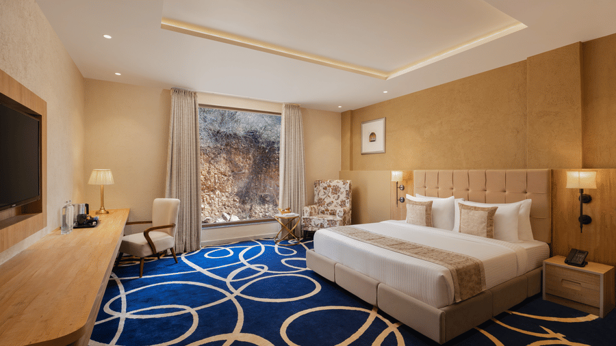 Deluxe room with a king size bed placed in front of a TV at Essentia Luxury Resort & Spa, Udaipur with a seating space by the window