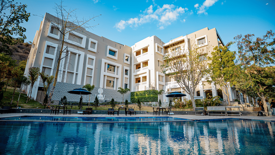 Facade view of Essentia Luxury Resort & Spa, Udaipur with a swimming pool in the foreground.